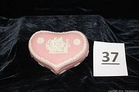 View of the pink heart-shaped Wedgwood trinket box against dark background showing top lid with white neo-classical relief design and rose border.