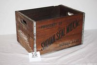 Rectangular wooden crate labeled 'PROPERTY OF SYLVAN SEAL MILK, Inc DEPOSIT CASE' with metal edge reinforcements and handle cutouts at sides, showing signs of usage and age.