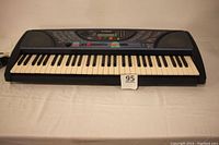 Front view of Yamaha PSR-248 keyboard showing full 61 keys, built-in speakers on left and right ends, and control panel on top center with various buttons and LCD screen. Keyboard is powered on as screen displays information.