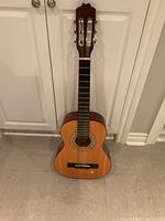 Full view of the half size classical acoustic guitar standing upright against a white door. The guitar has nylon strings and a traditional classical guitar headstock with open tuning pegs.
