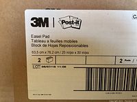 Close-up of product label on 3M Post-it easel pad showing brand, size 25 inch x 30 inch (63.5cm x 76.2cm), and lot information.