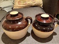 Two vintage ceramic crocks with lids shown from front on a gray couch surface, showcasing their two-tone brown and beige glossy glaze and intact lids.