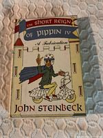 Front cover of John Steinbeck's The Short Reign of Pippin IV first edition hardcover showing colorful illustrated dust jacket with man on scooter and castle background.