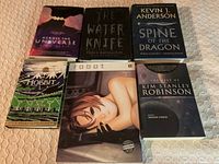 Photo showing the full lot of 6 books arranged on a flat surface, including Across the Universe, The Water Knife, Spine of the Dragon, The Hobbit, ⊙bot, and a Kim Stanley Robinson collection.