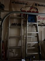 Photo of two ladders against a wall with banners showing brand logos including Honda, Suzuki, Ski-Doo, and EAO Sport. Left ladder is an extension type with curved top, right ladder is a step ladder with blue tray.