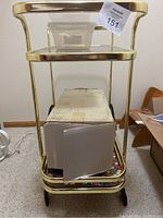 Full view of the gold metal bar cart showing the top and bottom glass shelves, the wheels, and the items stored on it, including plastic bins and a cardboard box.