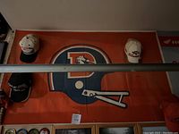 Denver Broncos orange flag with vintage helmet logo and four Broncos hats displayed on wall.