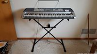 Full view of Casio keyboard on adjustable X-type metal stand against a wall, carpet flooring.