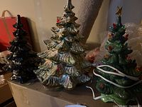 Photo showing all three ceramic Christmas trees placed side by side, dark colors and cream colored trees visible, on table in dim lighting.