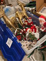 Plastic bin containing two gold-painted reindeer figurines, assorted Santa Claus figures, holiday decorations, and woven baskets.