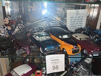 Black Corvette, purple Plymouth Prowler, metallic teal convertible, orange-black pickup, maroon convertible, partial view of motorcycles behind glass shelf
