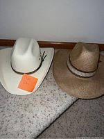 Two cowboy hats side by side on carpet floor, one white and one brown woven.