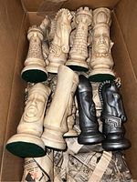 Photo showing 9 large giant chess pieces in light and dark colors packed in a cardboard box with newspaper cushioning, including rooks, knights, and kings.