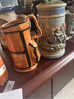 Photo showing barrel-shaped mug and motorcycle stein with detailed outdoor scene and figurine handle.