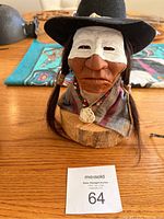 Close-up photo of Native American figurine with black hat, white face paint, and traditional shawl on wooden base.