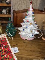 The two ceramic Christmas trees on a wooden table, one white and one green, visible tapestry edge.