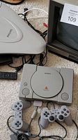 Photo showing Sony PlayStation console with two controllers and CRT TV in background.