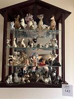 Front view of the wooden wall stand holding three glass shelves filled with a variety of ceramic cat figurines in different poses and colors.
