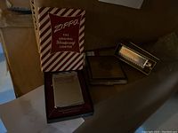 Photo showing a vintage Zippo lighter in original box, a gold-tone compact case, and a gold-tone lipstick tube all placed on a surface.