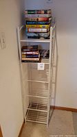 White wire shelving unit with six shelves, corner placement against wall, some books placed on top two shelves, unit generally clean but shows signs of dust and unclean surfaces