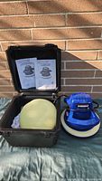 Simoniz polisher unit with foam pad on table beside open black plastic carrying case showing user manuals inside.