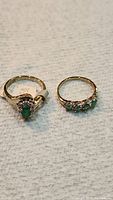 Two yellow gold electroplated rings with green stones laid side by side on a light surface.