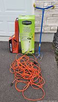 Overview photo of all items in the lot showing boxes of the Greenworks trimmer and Black & Decker hedge trimmer, blue garden weeding tool, and orange extension cord coiled.