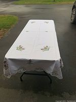 White rectangular table cloth with floral embroidery displayed on foldable black metal legs table outdoors on paved driveway.