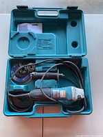 Open case showing Makita angle grinder, handle, grinding discs, nuts, washers, and manual inside.