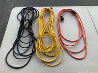 Photo showing three extension cords in black, yellow, and orange colors, coiled and bundled on a white table.