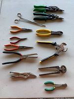 Photo of assorted hand tools including pliers, wire cutters, aviation snips, and locking pliers arranged on a beige surface.