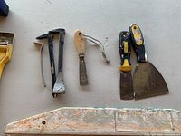 Top view of assorted drywall tools including two pry bars, wooden handled scraper, two metal putty knives, and metal razor blade scraper.