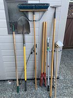 Photo showing full view of the assorted gardening tools including shovel, broom, and other long-handled tools leaning against a shed wall.