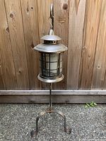 Front angle of the metal candle stand lantern showing cylindrical body with metal grid and glass panels, vented metal top, and three-legged base.