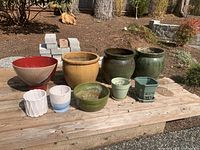 Photo showing 11 assorted ceramic pots arranged on wooden bench outdoors, varying in color, shape and size.