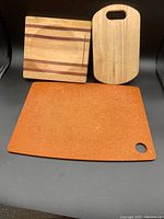 Photo showing three assorted cutting boards: one wooden with multi-tone stripes, one wooden with handle cut-out, and one brown synthetic with hanging hole.
