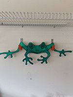 Front view of the green frog wall ornament mounted on a white wall under a wire shelf, showing the full width and outstretched legs and arms.