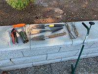 Set of assorted garden tools including pruners, trowels, cultivator, and bulb planter laid on concrete blocks.