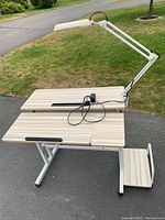 Front and side view of manual adjustable desk with attached desk lamp and footrest, showing wood grain surface, rubber edges, and silver metal base.