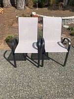 Two outdoor patio chairs placed side by side on a concrete ground, showing beige mesh fabric and black metal frames with armrests.