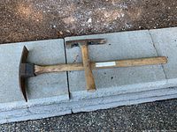 Two pick axes lying on concrete blocks outdoors on soil, showing heads and wooden handles with wear.