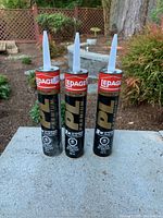Photo shows three unopened tubes of LEPAGE PL Premium Construction Adhesive standing upright outdoors with wooden fence and foliage in background.