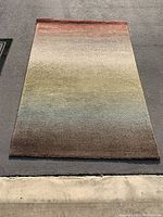Photo of a rectangular area rug with a horizontal gradient of colors including red, beige, green, and brown, laid out flat on a dark surface.