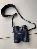 Top-side view of black Bushnell waterproof binoculars with neck strap laid on flat surface.