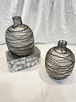 Two balloon-shaped glass vases with matte black and clear striping pattern on textured surface, shown against a white fabric backdrop, one vase slightly elevated on a block.