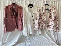 Three women's jackets hanging on black hangers against white fabric background, showing full front views of the mauve pink military jacket, white button-up coat, and floral blazer.
