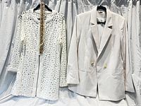 Photo of two women’s outerwear garments hanging against a curtain: a white cut-out patterned lightweight jacket/blouse and a white structured double-breasted blazer.