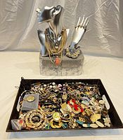 Silver-tone mannequins displaying layered necklaces, rings on hand, and pearl watch bracelet with a large tray full of assorted vintage style jewelry including earrings and brooches.