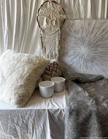 Photo showing macrame dream catcher with Tree of Life design hanging, framed sunburst wall art, two white ceramic pots, plush white and brown pillows, and gray faux fur throw blanket arranged on a white cloth surface.