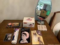Wide top-down view of lot on wooden table showing mirror, boxed LED nail lamp, eyeshadow palette, press-on nails, cotton pads, and hair caps.